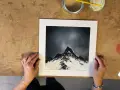 Des mains tiennent une œuvre d'art représentant des montagnes enneigées sous un ciel étoilé, rappelant le style des peintres contemporains.