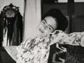 Frida Kahlo, le regard intense, pose dans une robe à volants. Derrière elle, un objet décoratif orné de franges, peut-être un indice sur son dernier tableau.