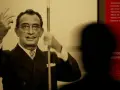Salvador Dalí, connu pour son surréalisme, pose avec ses mains levées. Un visiteur est en ombre chinoise devant un panneau rouge.