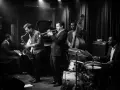 Un tableau musique noir et blanc d'un groupe de jazz jouant dans un club sombre. Un pianiste, un saxophoniste, un trompettiste, un contrebassiste et un batteur sont présents.