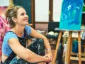 Jeune femme souriante dans son atelier, entourée de ses toiles. Elle pense à comment rechercher un artiste peintre pour sa prochaine acquisition.