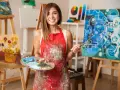 Une artiste peintre souriante, tablier taché de peinture, palette et pinceaux en main. Elle est entourée de ses œuvres, un studio qui inspire pour savoir comment devenir artiste peintre.