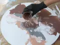 Main d'une personne versant de la peinture acrylique marron sur une toile ronde blanche, créant des motifs abstraits.