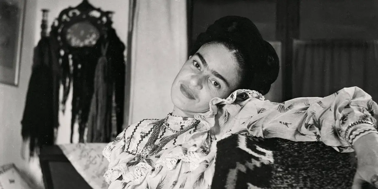 Frida Kahlo, le regard intense, pose dans une robe à volants. Derrière elle, un objet décoratif orné de franges, peut-être un indice sur son dernier tableau.