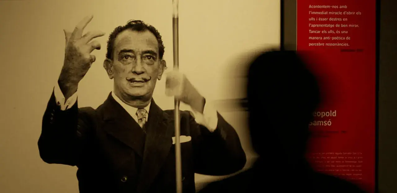 Salvador Dalí, connu pour son surréalisme, pose avec ses mains levées. Un visiteur est en ombre chinoise devant un panneau rouge.