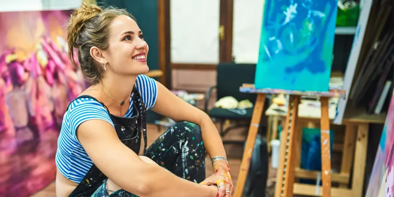Jeune femme souriante dans son atelier, entourée de ses toiles. Elle pense à comment rechercher un artiste peintre pour sa prochaine acquisition.