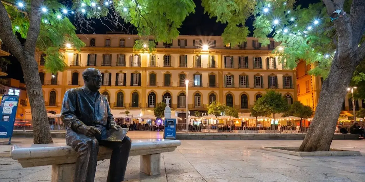 Statue de Picasso, rappelant ses origines, sur une place animée de Malaga la nuit, sous des arbres décorés de lumières.
