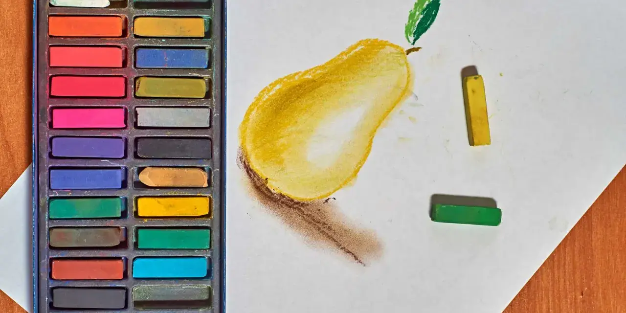 Une boîte de pastels secs pour débutant, avec une poire jaune esquissée sur du papier blanc, et des pastels jaune et vert à côté.