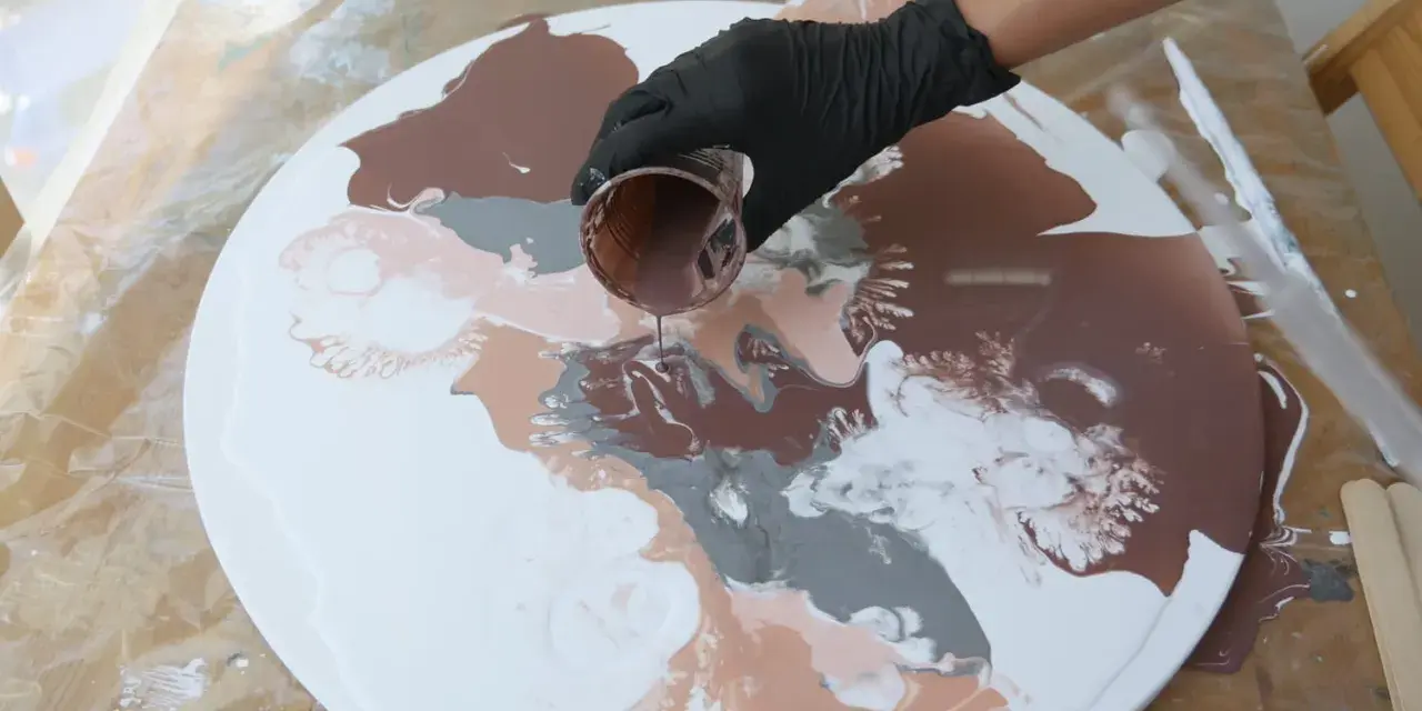 Main d'une personne versant de la peinture acrylique marron sur une toile ronde blanche, créant des motifs abstraits.