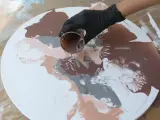 Main d'une personne versant de la peinture acrylique marron sur une toile ronde blanche, créant des motifs abstraits.