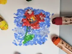 Apprenez comment utiliser les pastels à l'huile pour créer une fleur vibrante sur fond bleu. Des pastels jaune, rouge et orange sont visibles.