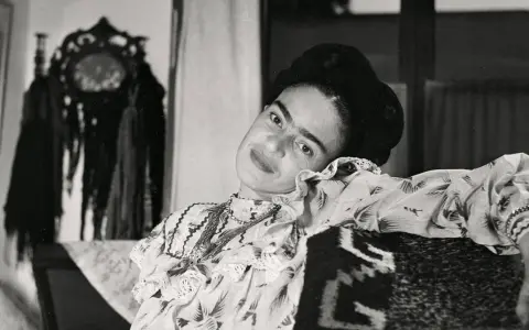 Frida Kahlo, le regard intense, pose dans une robe à volants. Derrière elle, un objet décoratif orné de franges, peut-être un indice sur son dernier tableau.