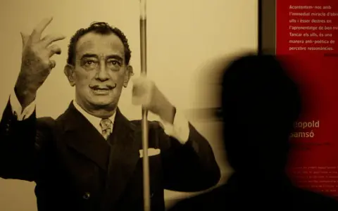 Salvador Dalí, connu pour son surréalisme, pose avec ses mains levées. Un visiteur est en ombre chinoise devant un panneau rouge.