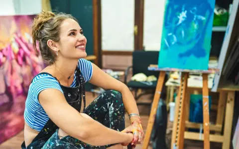Jeune femme souriante dans son atelier, entourée de ses toiles. Elle pense à comment rechercher un artiste peintre pour sa prochaine acquisition.