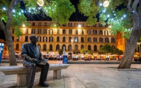 Statue de Picasso, rappelant ses origines, sur une place animée de Malaga la nuit, sous des arbres décorés de lumières.