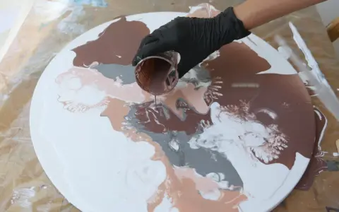 Main d'une personne versant de la peinture acrylique marron sur une toile ronde blanche, créant des motifs abstraits.