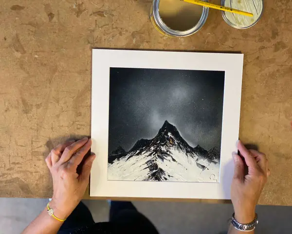 Des mains tiennent une œuvre d'art représentant des montagnes enneigées sous un ciel étoilé, rappelant le style des peintres contemporains.