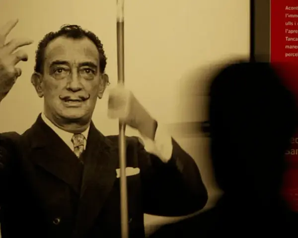 Salvador Dalí, connu pour son surréalisme, pose avec ses mains levées. Un visiteur est en ombre chinoise devant un panneau rouge.