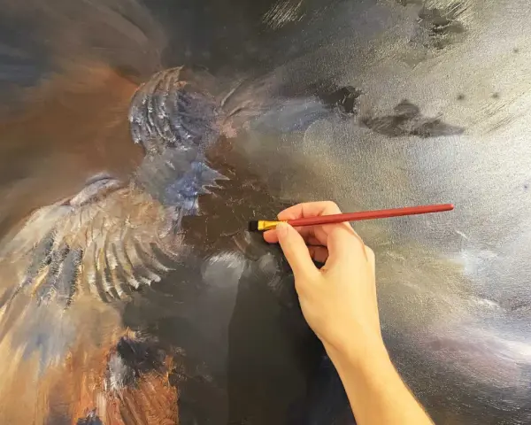 Main d'artiste travaillant sur une peinture à l'huile technique, ajoutant des détails à une figure aux ailes majestueuses.