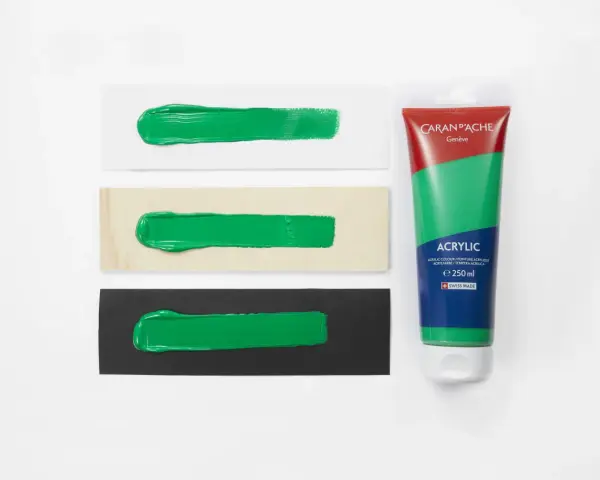 Bandeaux de peinture verte sur papier blanc, bois et noir, à côté d'un tube de gouache Caran d'Ache.