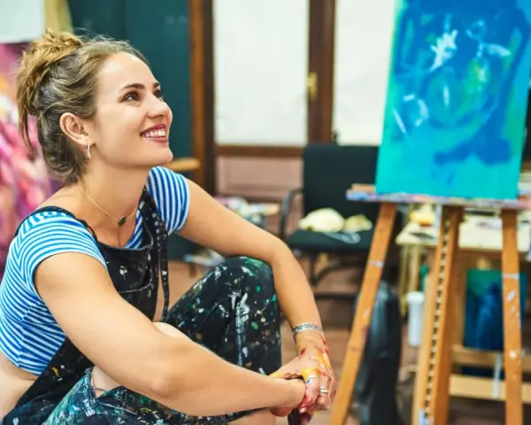 Jeune femme souriante dans son atelier, entourée de ses toiles. Elle pense à comment rechercher un artiste peintre pour sa prochaine acquisition.