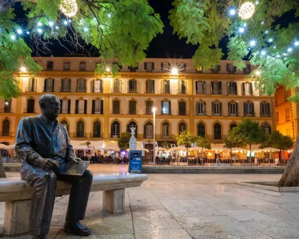 Statue de Picasso, rappelant ses origines, sur une place animée de Malaga la nuit, sous des arbres décorés de lumières.