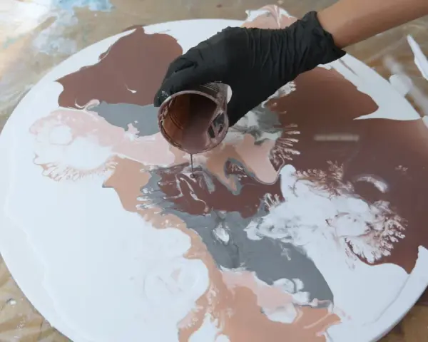 Main d'une personne versant de la peinture acrylique marron sur une toile ronde blanche, créant des motifs abstraits.