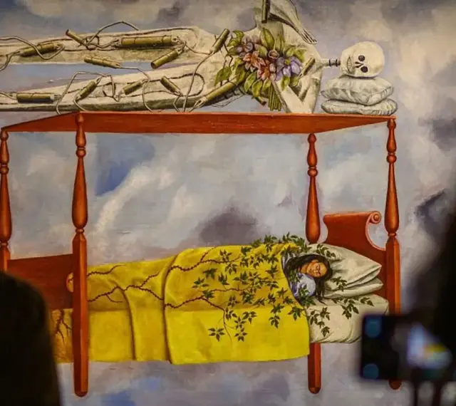 La peinture de Frida Kahlo "Le lit volant" montre une femme endormie sous une couverture jaune, avec un squelette au-dessus.