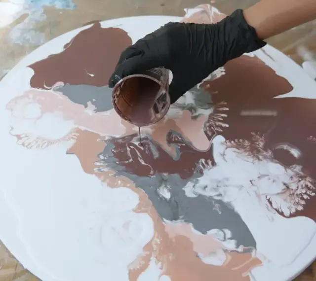 Main d'une personne versant de la peinture acrylique marron sur une toile ronde blanche, créant des motifs abstraits.