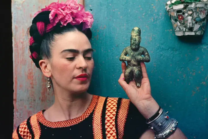 Frida Kahlo, avec ses fleurs roses et sa robe colorée, tient une statuette ancienne.