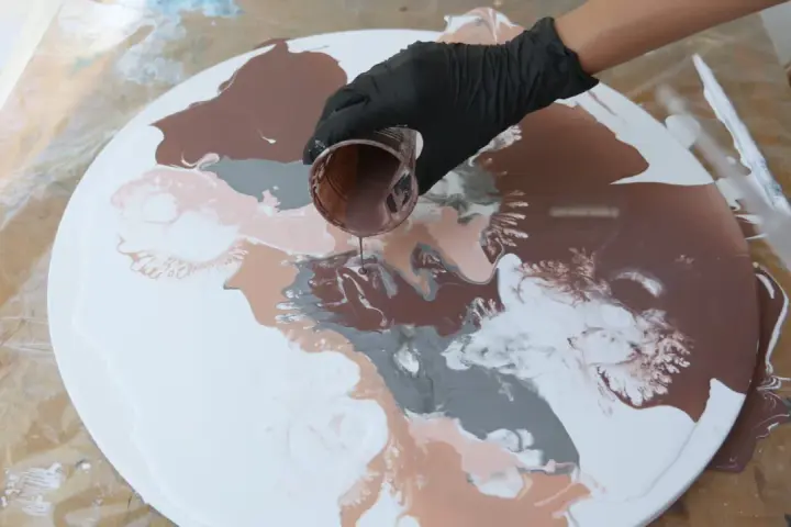 Main d'une personne versant de la peinture acrylique marron sur une toile ronde blanche, créant des motifs abstraits.