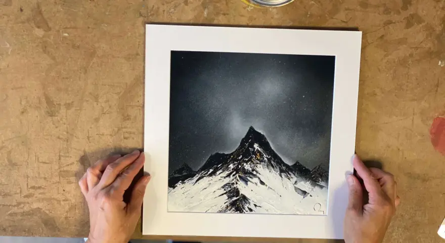 Des mains tiennent une œuvre d'art représentant des montagnes enneigées sous un ciel étoilé, rappelant le style des peintres contemporains.