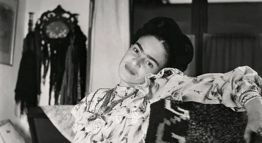 Frida Kahlo, le regard intense, pose dans une robe à volants. Derrière elle, un objet décoratif orné de franges, peut-être un indice sur son dernier tableau.
