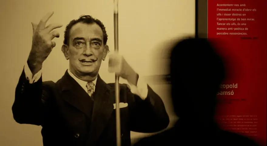 Salvador Dalí, connu pour son surréalisme, pose avec ses mains levées. Un visiteur est en ombre chinoise devant un panneau rouge.