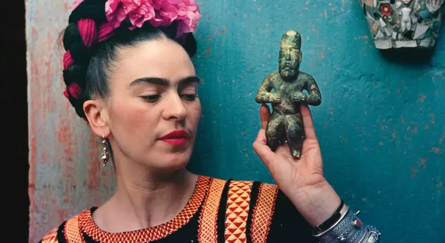 Frida Kahlo, avec ses fleurs roses et sa robe colorée, tient une statuette ancienne.