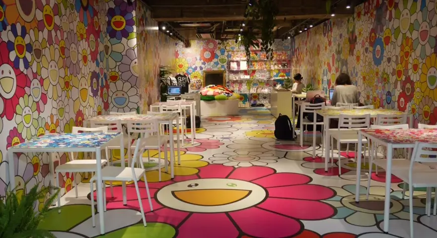 Un café aux couleurs vives, décoré de fleurs souriantes de l'artiste Murakami. Des tables et des chaises blanches invitent à s'asseoir dans cet univers joyeux.