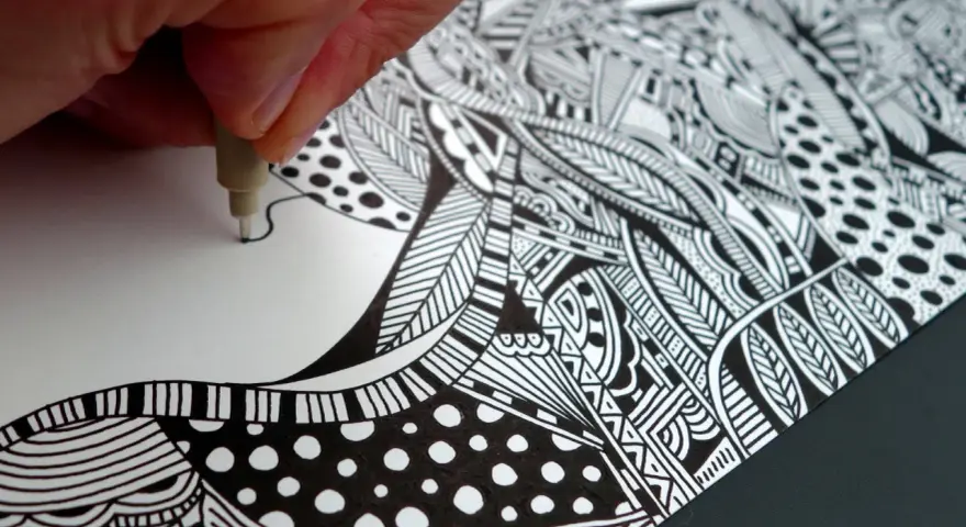 Main d'artiste créant un gribouillage artistique complexe, rempli de motifs géométriques et organiques en noir et blanc.