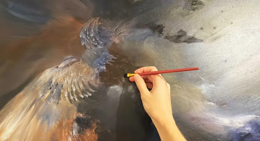 Main d'artiste travaillant sur une peinture à l'huile technique, ajoutant des détails à une figure aux ailes majestueuses.