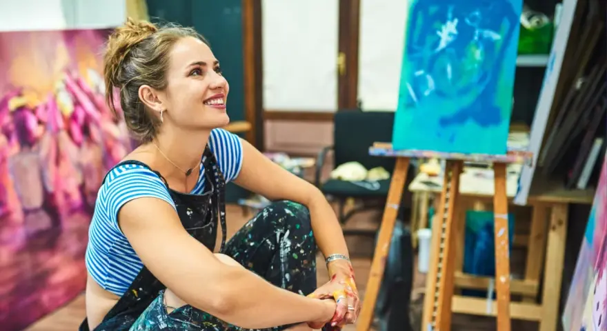 Jeune femme souriante dans son atelier, entourée de ses toiles. Elle pense à comment rechercher un artiste peintre pour sa prochaine acquisition.