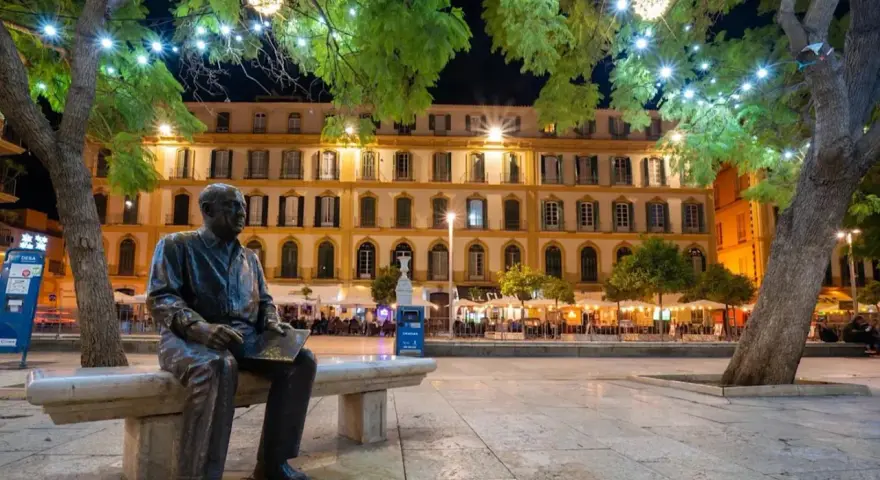 Statue de Picasso, rappelant ses origines, sur une place animée de Malaga la nuit, sous des arbres décorés de lumières.
