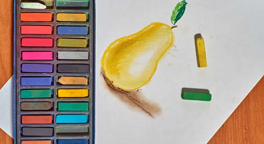 Une boîte de pastels secs pour débutant, avec une poire jaune esquissée sur du papier blanc, et des pastels jaune et vert à côté.