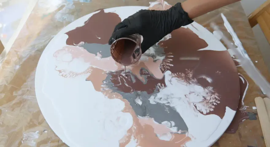 Main d'une personne versant de la peinture acrylique marron sur une toile ronde blanche, créant des motifs abstraits.
