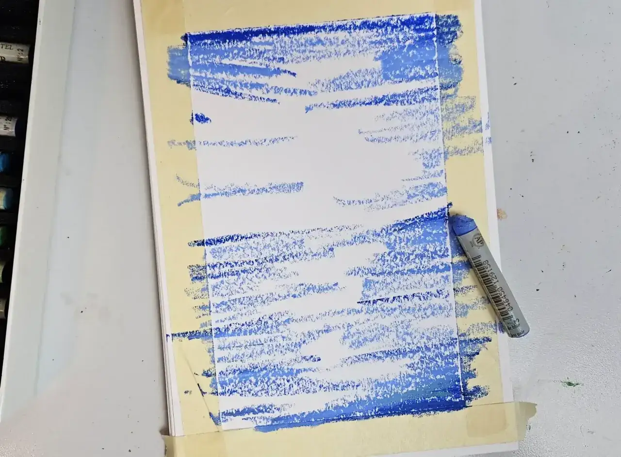D&eacute;couvrez comment utiliser les pastels &agrave; l'huile pour cr&eacute;er des effets de texture bleus sur un papier prot&eacute;g&eacute; par du masking tape. Un pastel bleu repose &agrave; c&ocirc;t&eacute;.