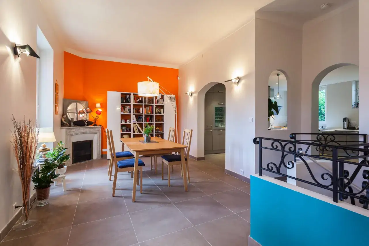 Un salon moderne avec un mur orange vif, une table en bois et des chaises bleues, contrastant avec des touches de bleu turquoise sur la balustrade.