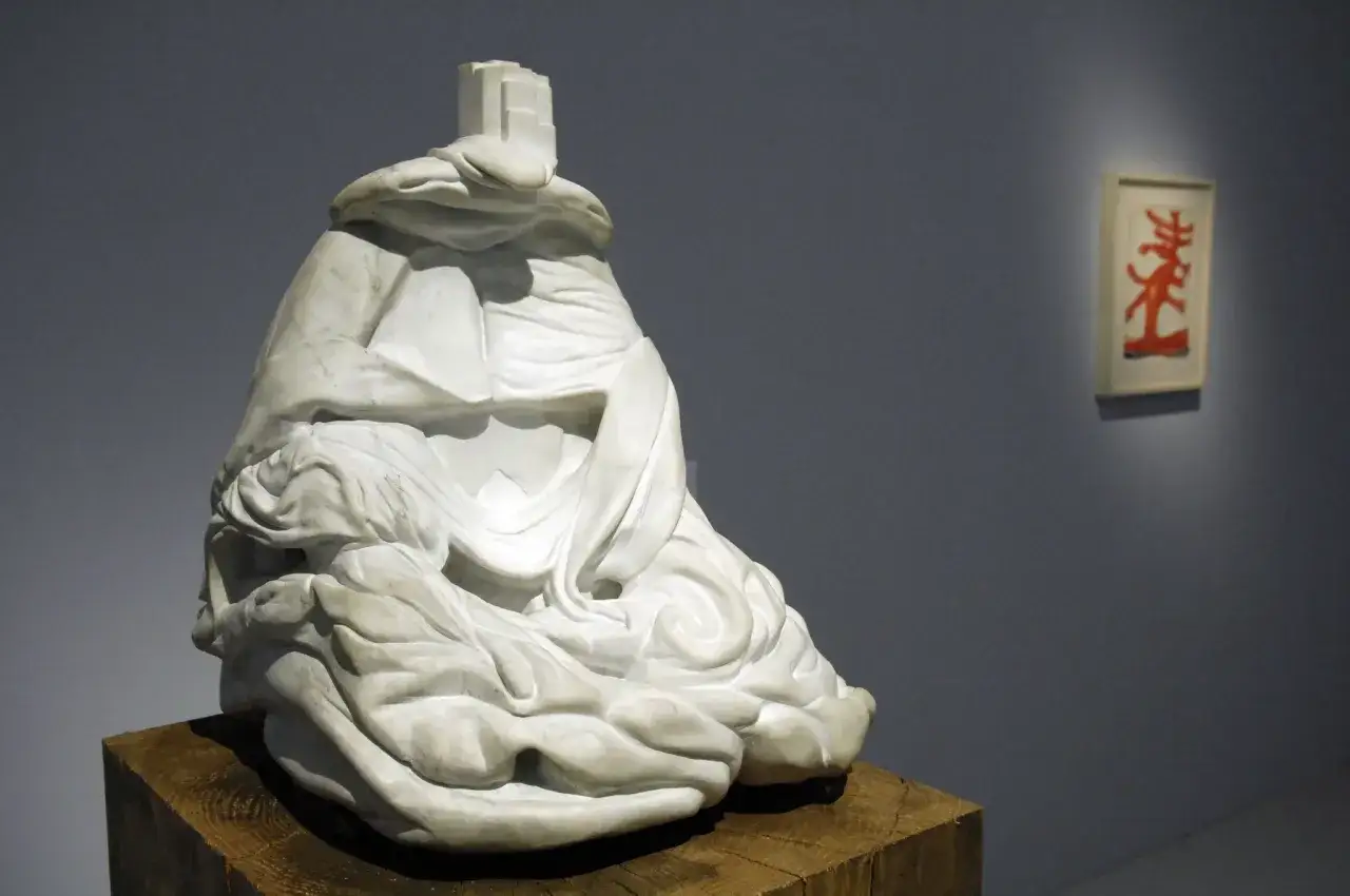 Sculpture en marbre blanc, une &oelig;uvre d'art de Louise Bourgeois, drap&eacute;e et assise sur un socle en bois. Une petite &oelig;uvre rouge est accroch&eacute;e au mur.
