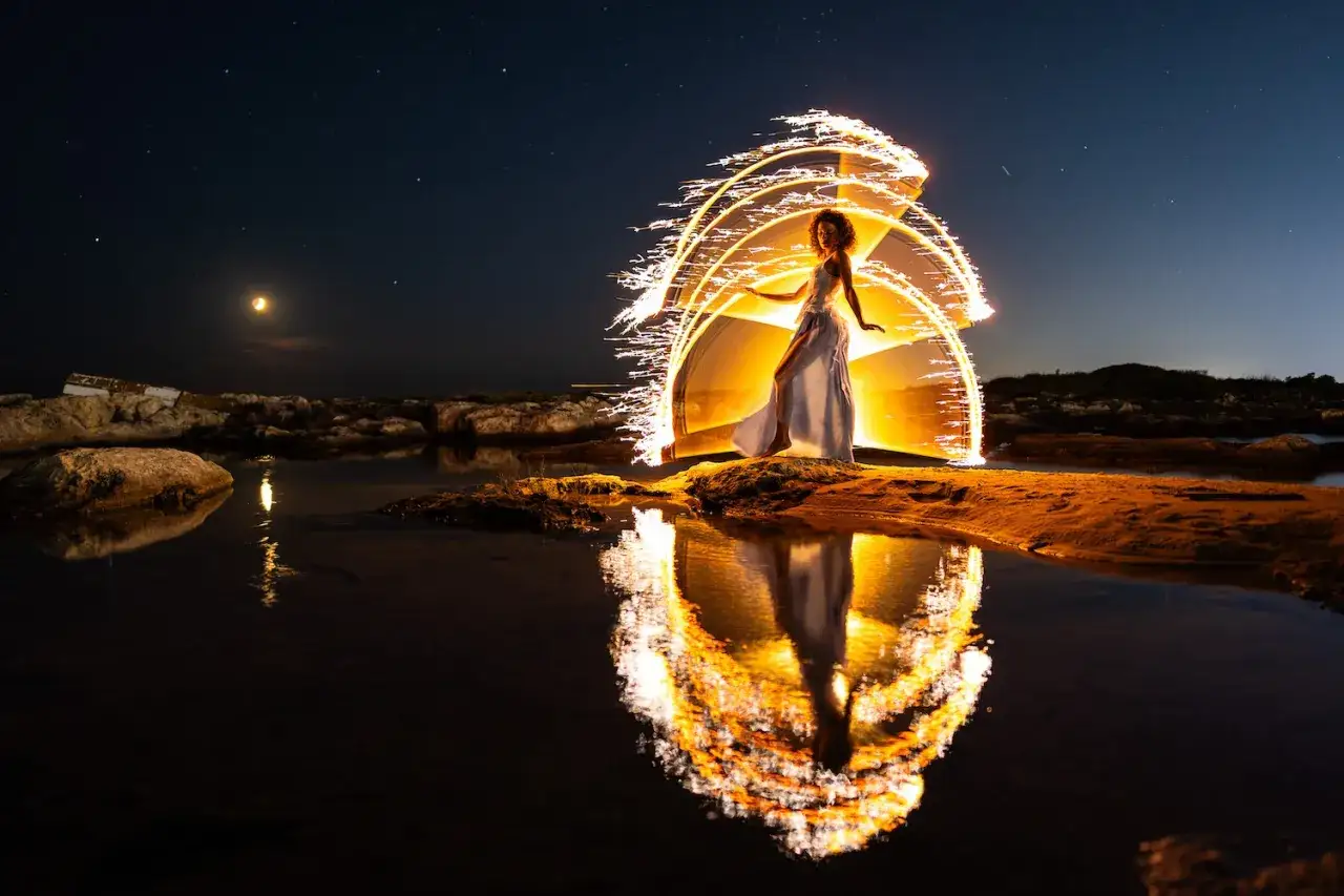 Une femme en robe blanche cr&eacute;e une peinture lumineuse la nuit, entour&eacute;e d'arcs de feu. Son reflet scintille dans l'eau sombre.